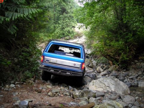 1985 Ford Bronco - A blue Ford Bronco is navigating a rocky, wooded trail, with the rear of the vehicle visible as it drives over stones and through a shallow…