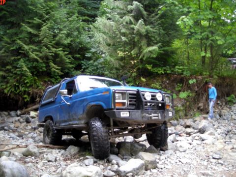 1985 Ford Bronco - A blue 1980s Ford Bronco is navigating a rocky, wooded terrain. A person stands to the right of the vehicle, observing its progress.