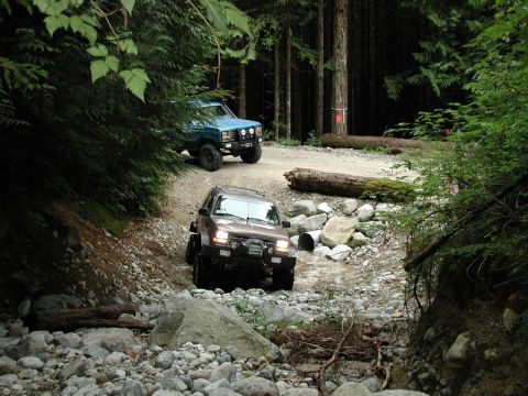 1985 Ford Bronco - Two off-road vehicles navigate a rocky, wooded trail. A brown SUV is in the foreground, crossing a shallow stream, while a blue pickup truc…