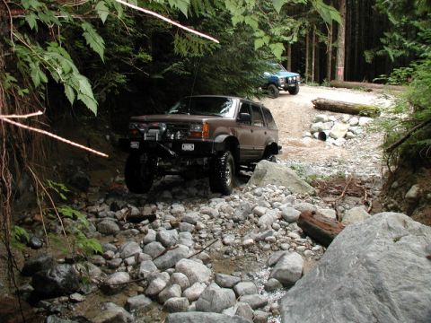1985 Ford Bronco - Two off-road SUVs navigate a rocky, wooded trail. The foreground vehicle, a brown SUV with off-road modifications, is positioned on a strea…