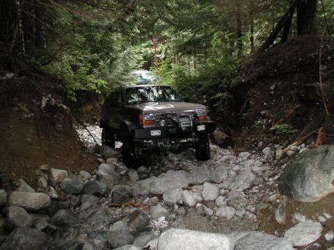1985 Ford Bronco - A lifted, dark-colored SUV navigates a rocky, wooded trail with a stream running alongside. Another vehicle is visible further up the trail.