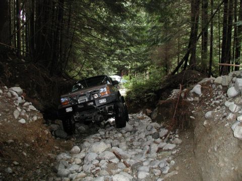 1985 Ford Bronco - A dark-colored SUV is navigating a rocky, wooded trail. The vehicle is angled to the left and appears to be driving through a shallow strea…