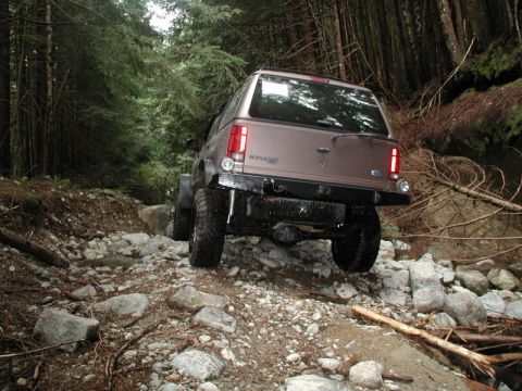 1985 Ford Bronco - A lifted, bronze-colored SUV navigates a rocky, wooded trail. The rear of the vehicle is visible, showing large off-road tires and a custom…