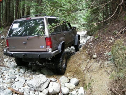 1985 Ford Bronco - The rear of a brown Ford Explorer SUV is shown navigating a rocky, wooded trail. The vehicle has a custom rear bumper and is positioned on…