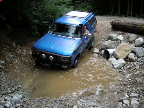 1985 Ford Bronco - A blue Ford Bronco with a camper shell is driving through a muddy puddle on a dirt road, with a large log and rocks on the side of the road.