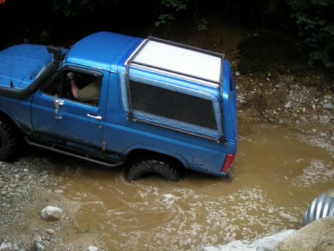 1985 Ford Bronco - A blue Ford Bronco is driving through a shallow, muddy stream. The vehicle has a snorkel intake on the driver's side and a white camper she…