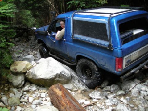 1985 Ford Bronco - A blue 1980s Ford Bronco is navigating a rocky, wooded trail. The vehicle is positioned at an angle, with its right side visible as it driv…