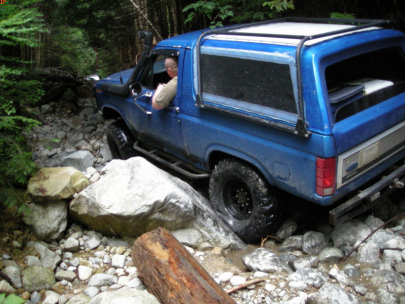 Blue 1980s Ford Bronco off-roading on a rocky, wooded trail.