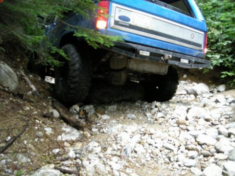 1985 Ford Bronco - The rear passenger side of a blue Ford Bronco is shown navigating a rocky, wooded trail. The vehicle's large off-road tire is visible, as i…