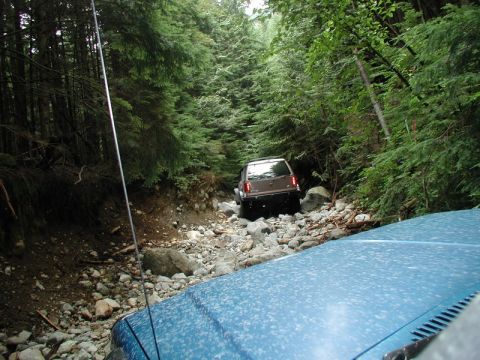 1985 Ford Bronco - A blue vehicle's hood is in the foreground, looking out at a brown SUV navigating a rocky, wooded trail. The SUV is positioned further up t…