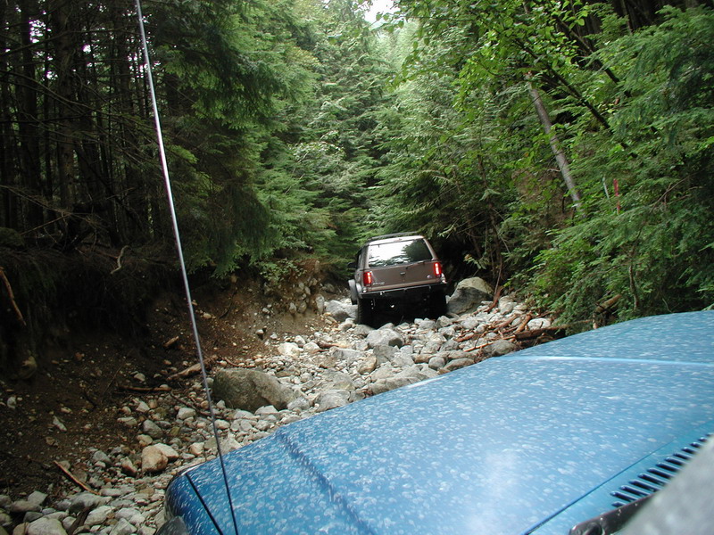 Blue vehicle hood in foreground, looking at brown SUV on a rocky forest trail.
