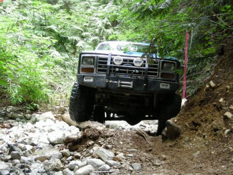 1985 Ford Bronco - A blue Ford Bronco is navigating a rocky, wooded trail, with its front tires angled towards the right side of the frame. The vehicle has an…