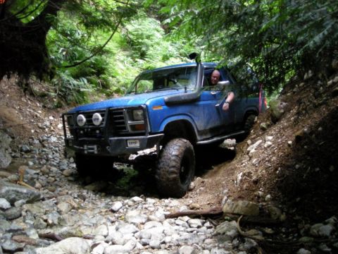 1985 Ford Bronco - A blue Ford Bronco is navigating a rocky, wooded trail with a driver visible behind the wheel. The vehicle has a snorkel, a custom bumper w…