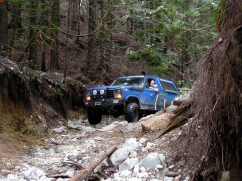 1985 Ford Bronco - A blue Ford Bronco is navigating a rocky, wooded trail. The vehicle is equipped with off-road tires, a snorkel, and auxiliary lights on the…