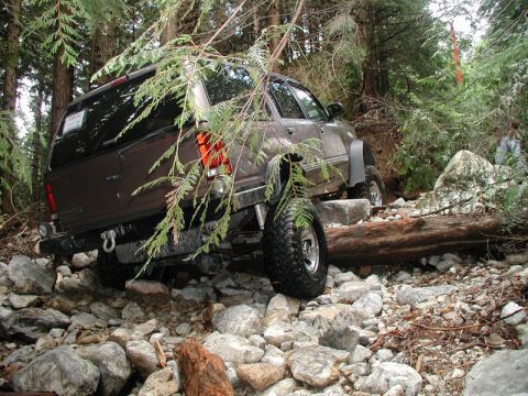 1985 Ford Bronco - A brown SUV is navigating a rocky, wooded trail, with its rear tire lifted over a large rock and a fallen log in the foreground.