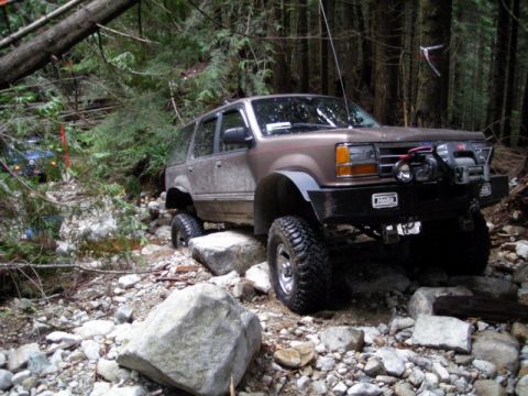 1985 Ford Bronco - A lifted, brown Ford Explorer is navigating a rocky, wooded trail, with its front tire resting on a large rock. A blue SUV is visible furth…