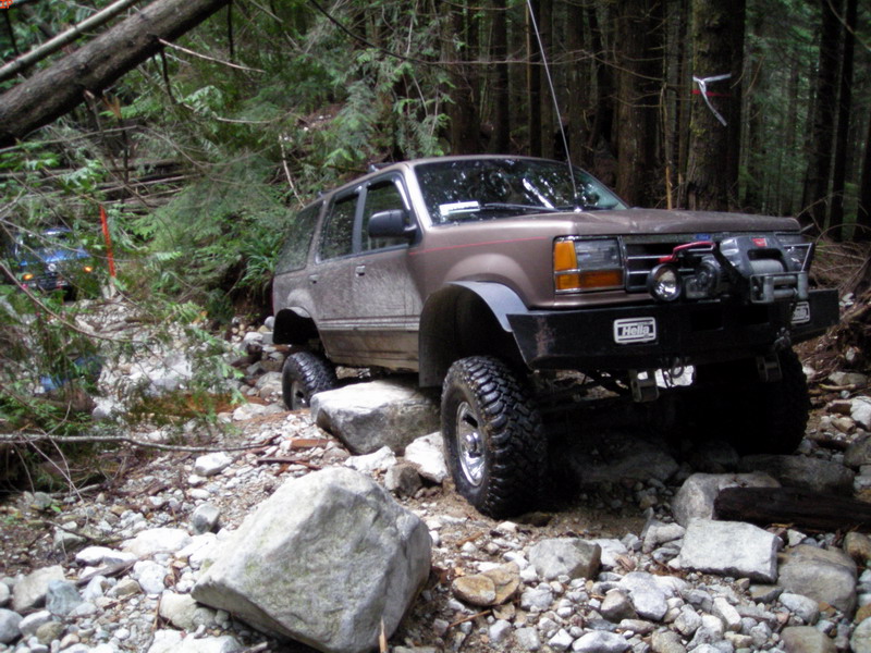 Lifted brown Ford Explorer off-roading on a rocky, wooded trail.
