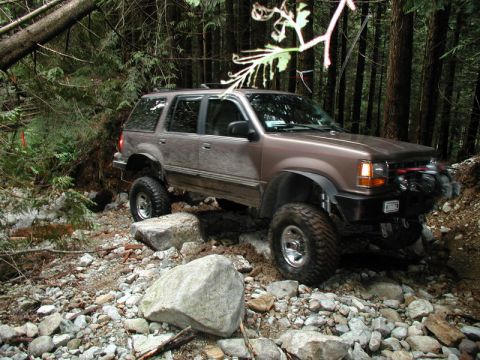1985 Ford Bronco - A lifted, brown SUV is navigating a rocky, wooded trail. The vehicle is angled, with its front right tire high on a rock and its front bump…