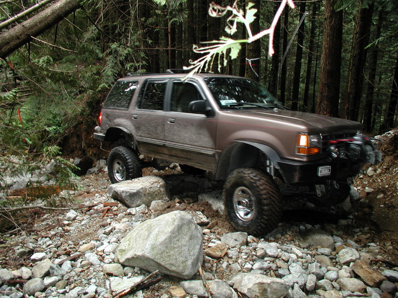 Lifted brown SUV with winch on a rocky, wooded trail.