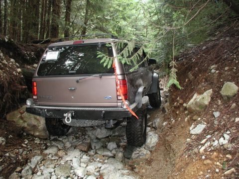 1985 Ford Bronco - A tan Ford Explorer is navigating a rocky, wooded trail, with its rear tire on a large rock. An orange strap is visible on the rear of the…