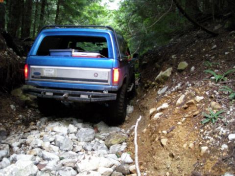 1985 Ford Bronco - The rear of a blue Ford Bronco is navigating a rocky, wooded trail. The vehicle is positioned on a steep incline with loose rocks and dirt…