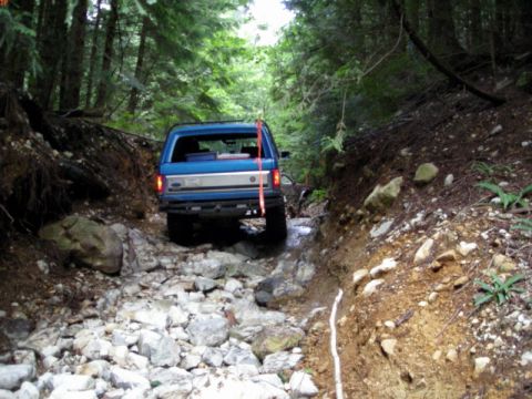 1985 Ford Bronco - A blue Ford Bronco is navigating a rocky, off-road trail through a dense forest. The vehicle is viewed from the rear as it drives over a st…