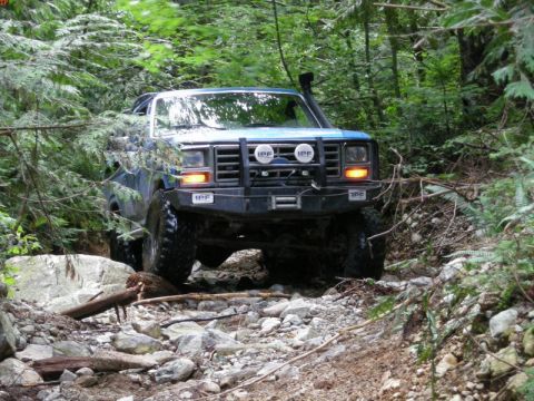 1985 Ford Bronco - A blue Ford Bronco navigates a rocky, wooded trail, its front end visible with off-road lights and a snorkel.