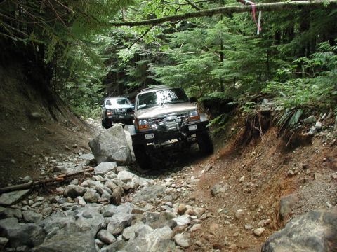 1985 Ford Bronco - Two off-road vehicles navigate a rocky, wooded trail. The lead vehicle, a tan SUV, is angled towards the right side of the frame, showcasin…