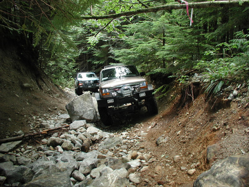 Two off-road SUVs on a rocky, wooded trail.