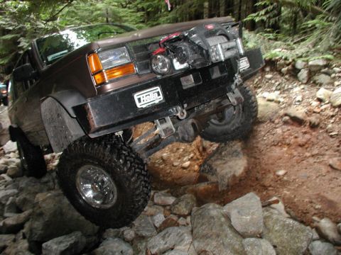1985 Ford Bronco - A brown Ford Bronco is navigating a rocky, off-road trail. The vehicle has large tires, a custom bumper with a winch, and a "Hella" sticker…