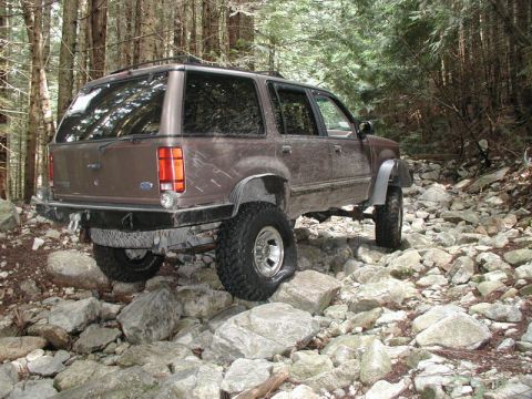 1985 Ford Bronco - A brown, lifted SUV navigates a rocky, wooded trail. The vehicle has large off-road tires and a modified rear bumper.