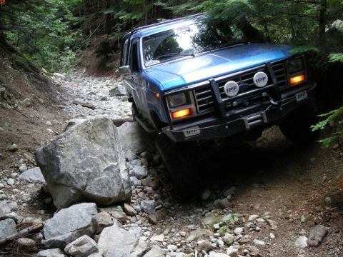 1985 Ford Bronco - A blue 1985 Ford Bronco is navigating a rocky, wooded trail, with its front tire climbing over a large boulder. The vehicle has aftermarket…