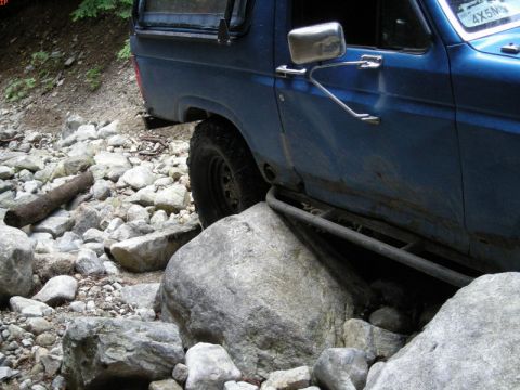 1985 Ford Bronco - The side of a blue Ford Bronco is shown navigating a rocky, off-road terrain. The vehicle has aftermarket rock sliders installed and is pos…
