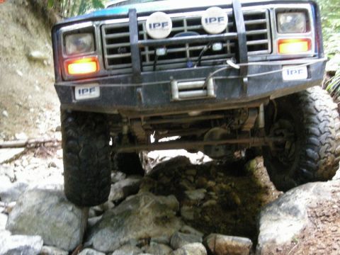 1985 Ford Bronco - The front of a blue off-road vehicle is shown navigating a rocky trail. The vehicle has a custom bumper with a winch and IPF lights, and th…