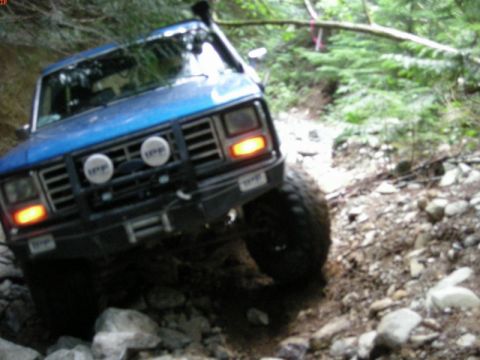 1985 Ford Bronco - A blue Ford Bronco is navigating a rocky, wooded trail. The front of the SUV is visible, featuring auxiliary lights and a snorkel.