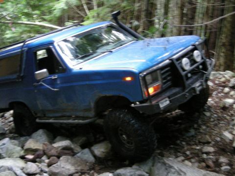 1985 Ford Bronco - A blue Ford Bronco is navigating a rocky, wooded trail. The vehicle has large off-road tires, a snorkel, and a brush guard with auxiliary l…