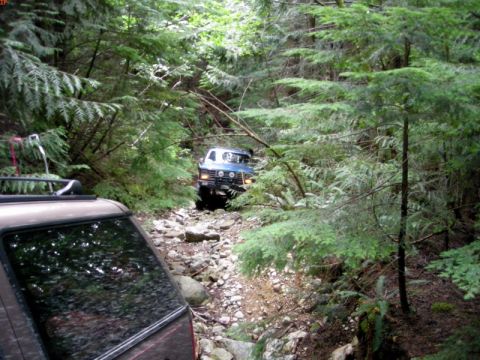 1985 Ford Bronco - A blue 1980s SUV is navigating a rocky, wooded trail. The rear of a brown SUV is visible in the foreground.