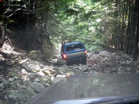 1985 Ford Bronco - A blue Ford Bronco is navigating a rocky, wooded trail. The photo is taken from the perspective of another vehicle following behind.