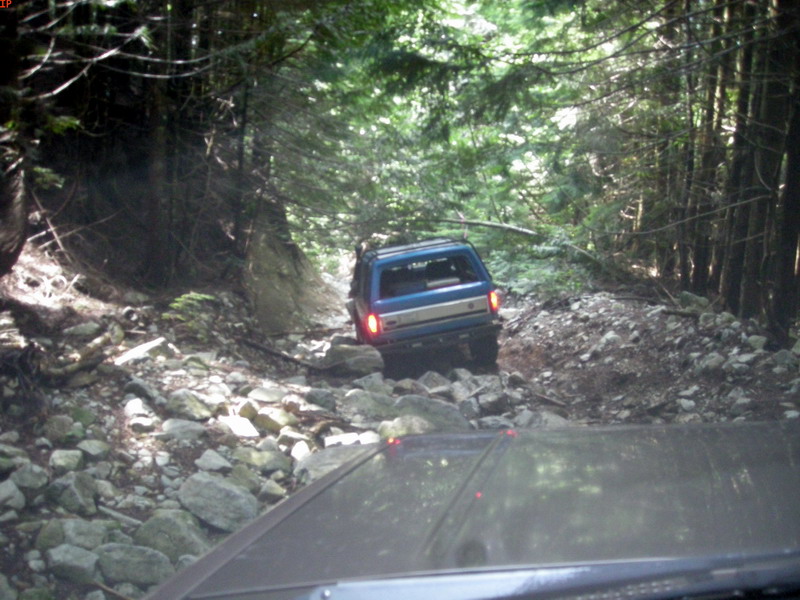 Blue Ford Bronco driving on a rocky, wooded trail.