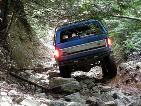 1985 Ford Bronco - A blue Ford Bronco is navigating a rocky, wooded trail. The rear of the SUV is visible as it climbs over obstacles.