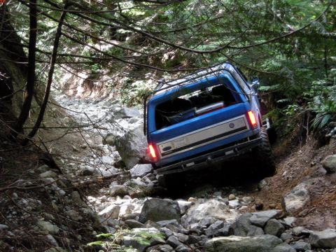 1985 Ford Bronco - A blue Ford Bronco is navigating a steep, rocky trail in a dense forest. The vehicle is angled to the right, with its rear facing the camer…
