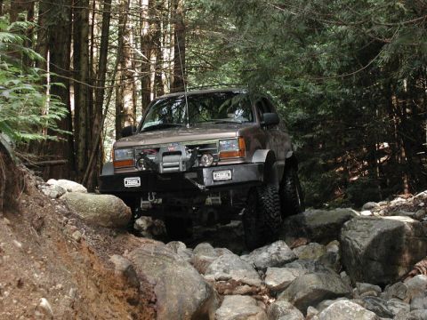 1985 Ford Bronco - A brown off-road SUV navigates a rocky, wooded trail. The vehicle features a winch and a custom bumper with auxiliary lights.
