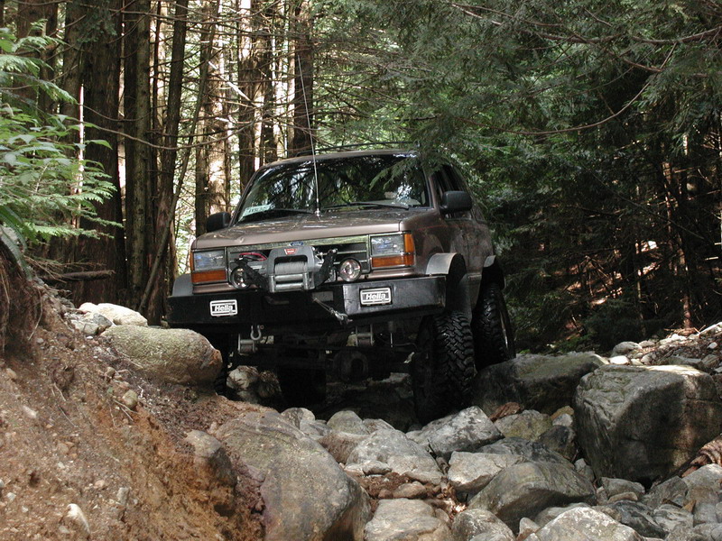 Brown off-road SUV on a rocky trail in a forest, featuring a winch and custom bumper.