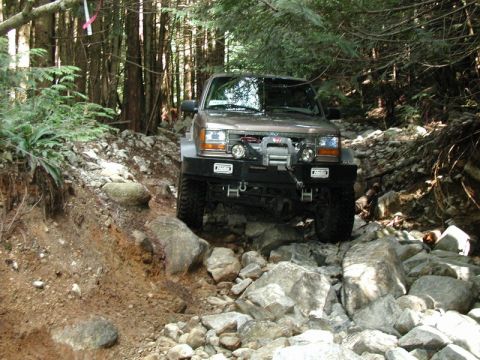 1985 Ford Bronco - A brown 1985 Ford Bronco is navigating a rocky, wooded trail. The vehicle is equipped with a winch and aftermarket bumper.