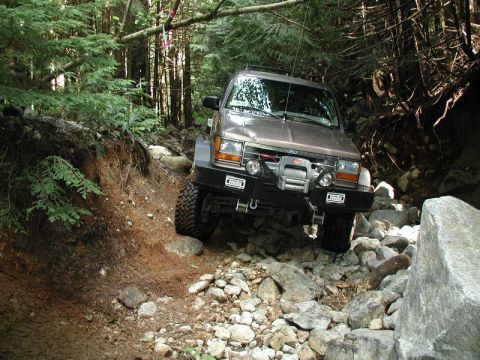 1985 Ford Bronco - A brown Ford Explorer is navigating a rocky, wooded trail, with its front tires on a steep incline and its winch visible on the aftermarket…