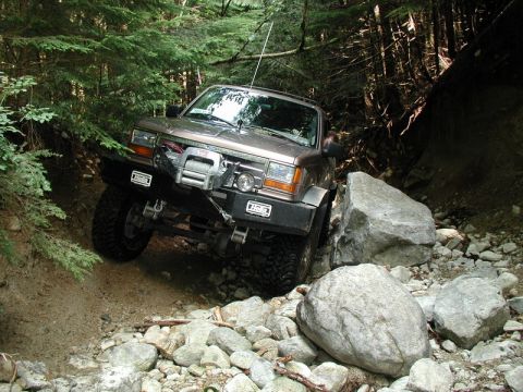 1985 Ford Bronco - A brown 1980s Ford Bronco is navigating a rocky, wooded trail, with its front tire angled up against a large boulder. The vehicle is equipp…