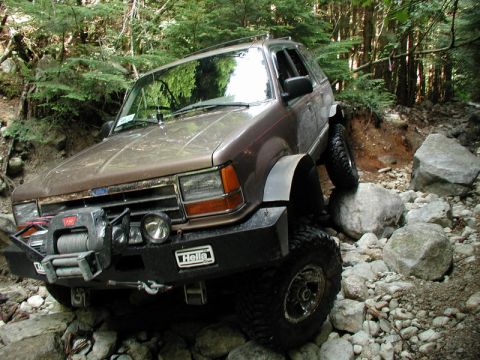 1985 Ford Bronco - A brown Ford Explorer is navigating a rocky, wooded trail, with its front tire on top of a large boulder. The vehicle is equipped with a wi…