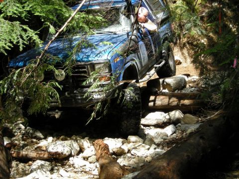 1985 Ford Bronco - A blue 1980s Ford Bronco is navigating a rocky, wooded trail, with its front tires on wooden logs. The vehicle is surrounded by trees and r…