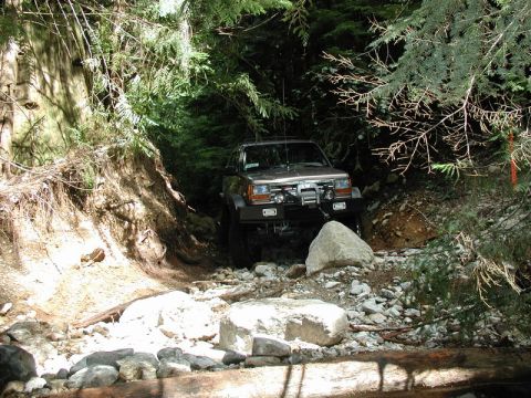 1985 Ford Bronco - A brown SUV navigates a rocky, wooded trail, with a winch mounted on the front bumper. The vehicle is surrounded by dense trees and large r…