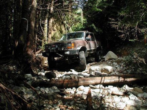 1985 Ford Bronco - A brown Ford Explorer is navigating a rocky, wooded trail. The SUV is equipped with off-road tires and auxiliary lights on the front bumper.